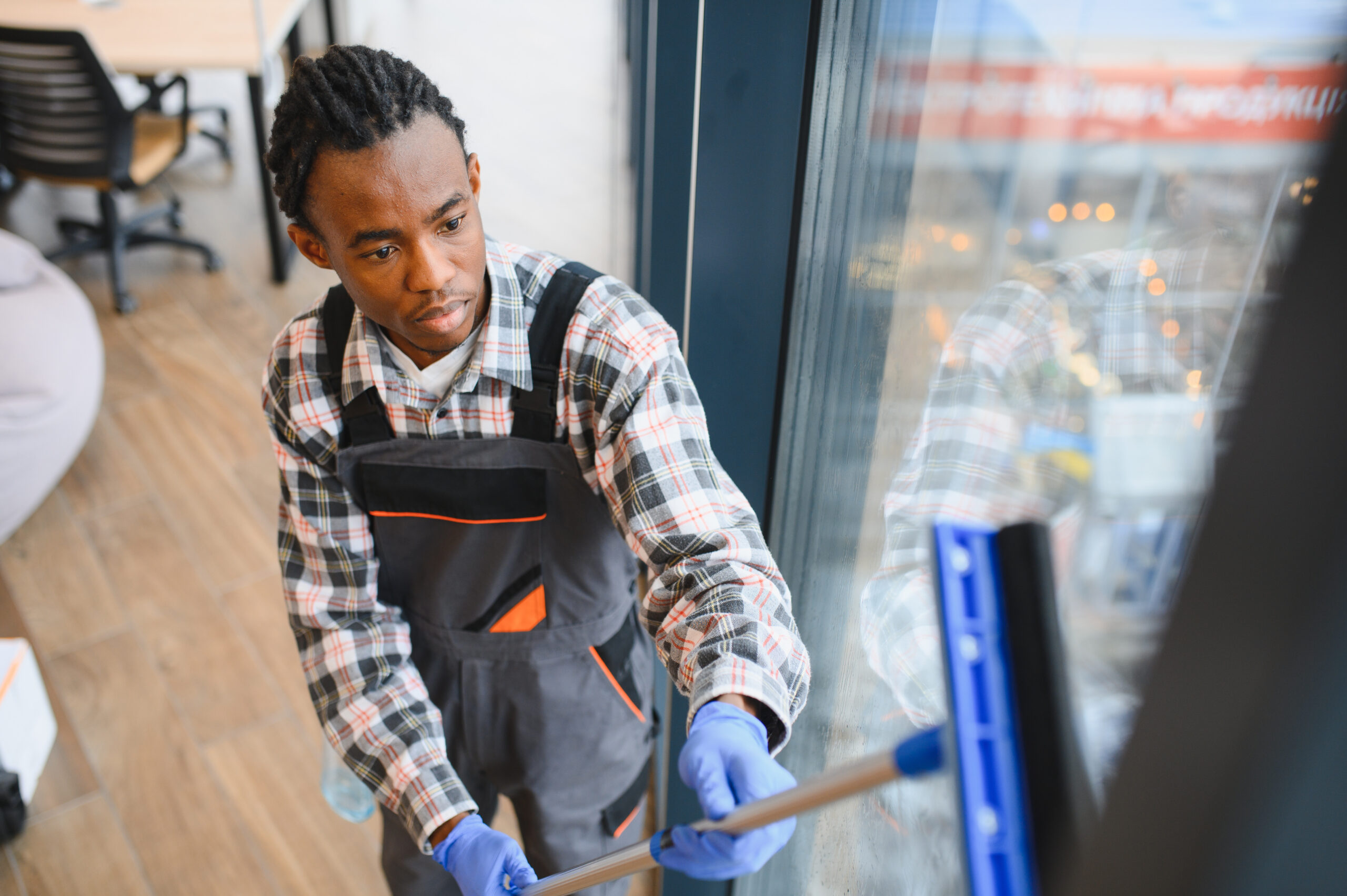 Young african american male janitor wearing uniform and gloves cleaning windows with squeegee in office building