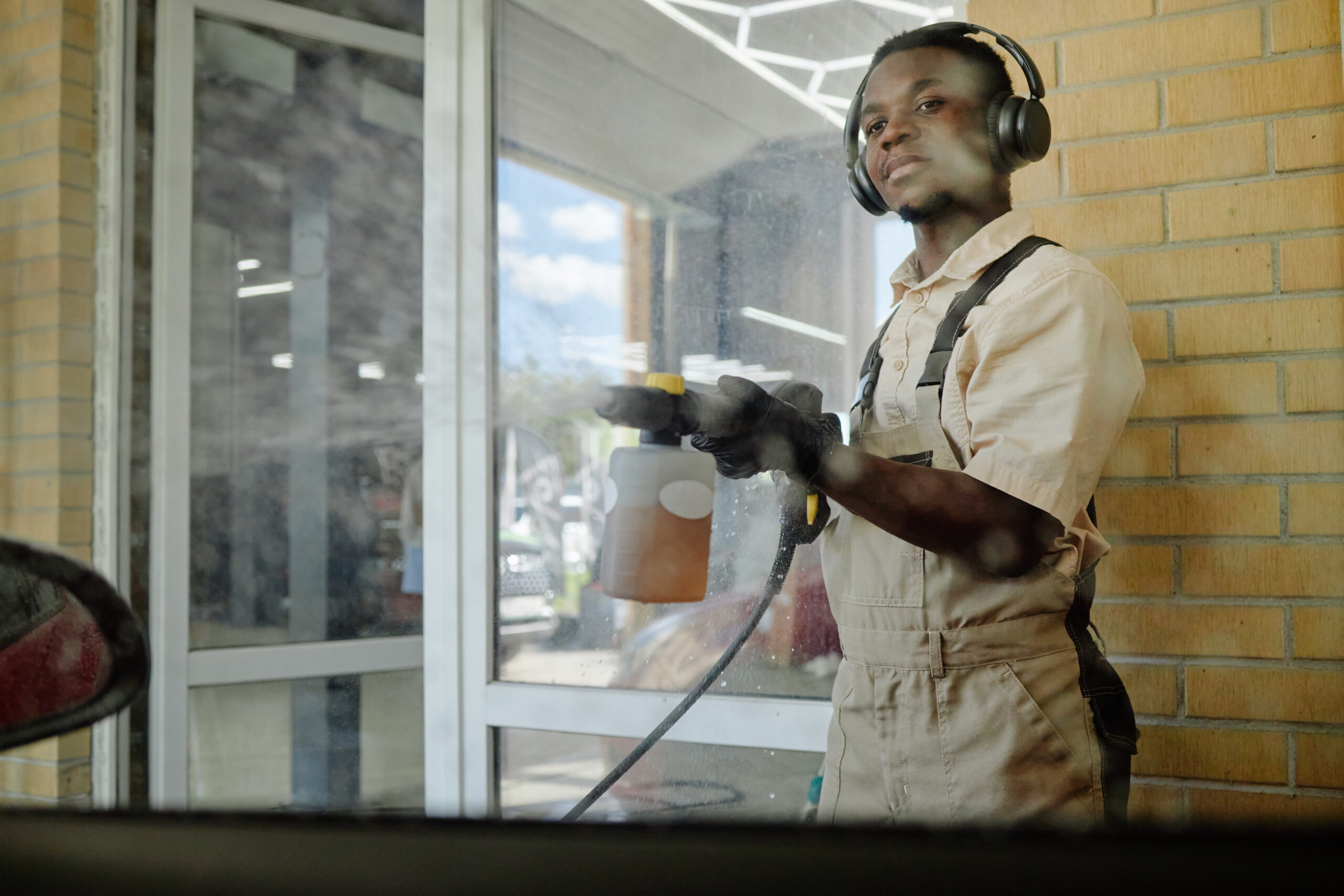 Young Man Wearing Headphones Cleaning Automobile with Professional Equipment Young adult Black man wearing headphones cleaning automobile with high pressure washer in garage